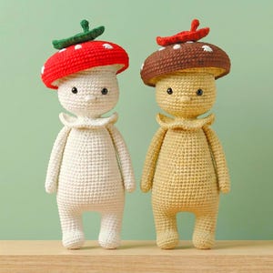 Puede incluir: Dos muñecos de setas de ganchillo. Un muñeco es de color blanco roto con una gorra de seta roja con lunares blancos y un tallo verde. El otro muñeco es amarillo con una gorra de seta marrón con lunares blancos y un tallo rojo. Ambos muñecos tienen un cuello con volantes.