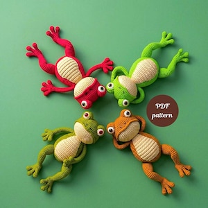 May include: Four crocheted frog toys in red, green, and orange, with cream-colored bellies. The frogs are arranged in a circle on a green background. The text "PDF pattern" is in a brown circle.