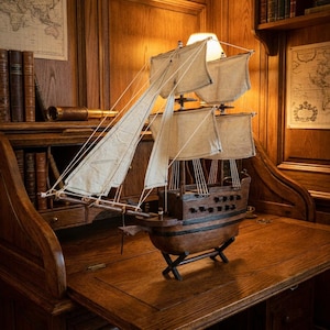 May include: A wooden model ship featuring a dark brown hull with black horizontal detailing, multiple masts, and off-white fabric sails rigged with thin white cordage. The vessel is supported by a black X-shaped stand on a flat wooden surface.
