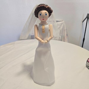 May include: A wooden doll dressed as a bride, featuring a white gown, a sheer white veil, and brown hair. The doll has painted blue eyes, red lips, and wears a silver-toned necklace and bracelet. The figure stands upright with arms positioned forward.