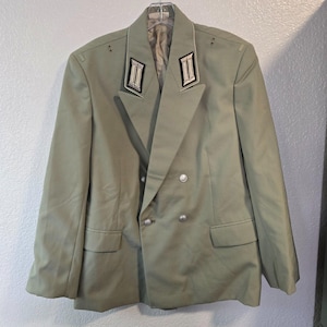 May include: A light olive green double-breasted jacket featuring silver-toned buttons and black and silver collar tabs with rectangular detailing. The garment has a notched lapel, two front flap pockets, and shoulder epaulets, presented on a hanger.