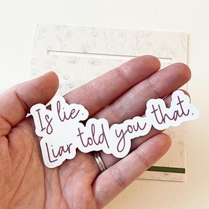 Può includere: Un adesivo bianco con il testo "Is lie. Liar told you that" in uno script viola scuro. L'adesivo è tenuto in mano, con una carta a motivi floreali sullo sfondo.