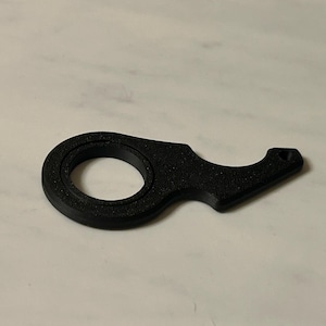 May include: A black, 3D-printed bottle opener with a ring handle and a curved lever. The opener is made of a textured material and is designed for easy gripping. The overall design is simple and functional.