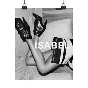 May include: Black and white fashion print featuring the word "ISABEL" in large white letters. The image shows a person wearing black high-heeled shoes with multiple straps and buckles. A black and white striped garment is also visible.
