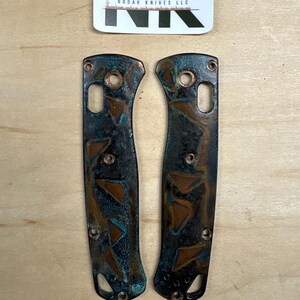 Peut inclure: Deux plaquettes de manche de couteau avec une finition cuivrée et sarcelle vieillie. Les plaquettes présentent un motif géométrique et sont étiquetées "Ball Python". Une étiquette avec "NODAK KNIVES LLC" est en haut.
