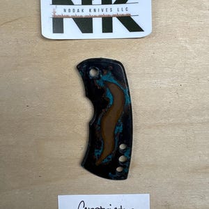Könnte beinhalten: Ein Messergriff mit schwarzer und türkisfarbener Oberfläche, mit einer einzigartigen Form und drei Bohrungen. Der Griff ist mit "Constrictor" beschriftet und wird von einem "Nodak Knives LLC" Aufkleber begleitet.
