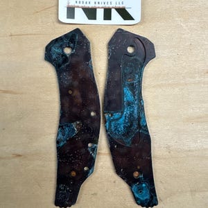 May include: Two weathered metal knife scales with a blue-green patina. The scales are irregularly shaped with several holes and a dark brown base color. A sticker with the text "Nodak Knives LLC" is above the scales, and a label that says "Jungle Gym" is below.