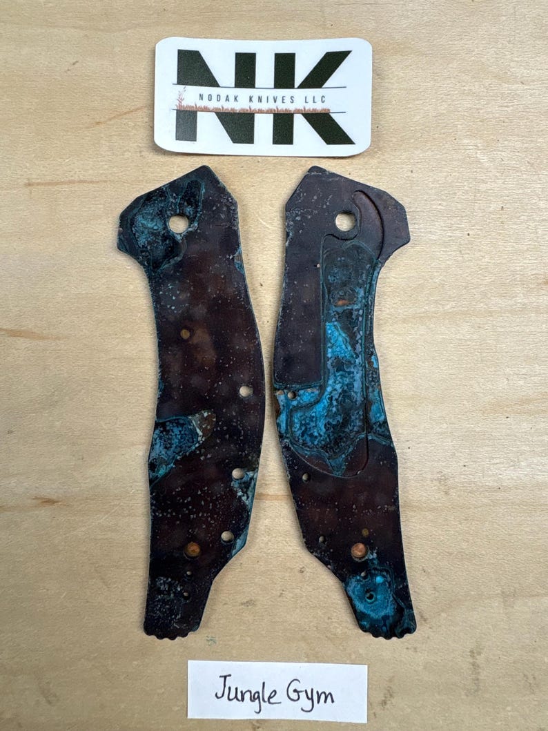 May include: Two weathered metal knife scales with a blue-green patina. The scales are irregularly shaped with several holes and a dark brown base color. A sticker with the text "Nodak Knives LLC" is above the scales, and a label that says "Jungle Gym" is below.