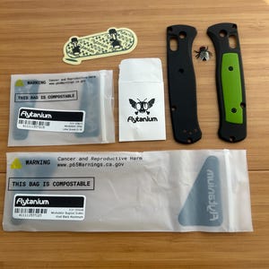 May include: A set of Flytanium knife parts including two black handle scales with a lime green inlay, a small fly figurine, a branded sticker, and two compostable plastic bags with warning labels and product information for Modulator Bugout scales.
