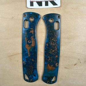 May include: Two knife handle scales featuring a mottled blue and copper patina finish. The scales have pre-drilled holes for assembly. A white label above reads NK NODAK KNIVES LLC and a white label below reads Captain Hook in black text.