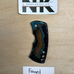 Op de afbeelding: Een meshandvat met een zwart, blauwgroen en bruin ontwerp. De handgreep heeft drie gaten en is gelabeld "Fanged". Het logo "NK" en "NODAK KNIVES LLC" staat boven de handgreep.