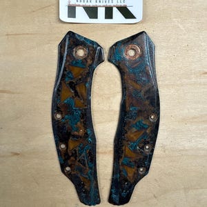 May include: Two knife handle scales with a brown and teal patina. The scales have multiple holes for attachment and a copper rivet. The top of the image has a sticker with the text "NK" and "NODAK KNIVES LLC". The bottom of the image has the text "Jungle Gym".