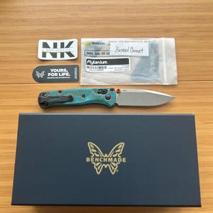 Op de afbeelding: Een teal en zilveren vouwmes met een zilveren lemmet, een zwarte doos met het Benchmade-logo en stickers met de tekst "NK" en "Yours, for Life". Op een zak staat "Rusted Comet".