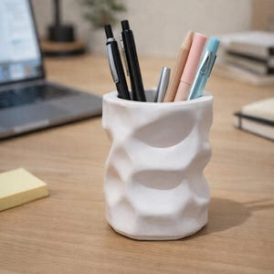 May include: A white cylindrical pen holder with a textured, faceted surface resembling crumpled paper. The holder contains a collection of writing instruments, including black pens, a silver pen, a wooden pencil, a pink pen, and a light blue pen.