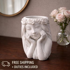 May include: A white ceramic planter sculpted in the form of a woman's face, adorned with a floral crown. The woman's hands are delicately placed on her cheeks. The planter sits on a dark wooden surface.