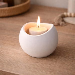 May include: A matte white ceramic candle holder with a rounded, bowl-like shape. The holder features a smooth, curved exterior and a wide opening at the top, containing a single lit tea light candle with a visible flame and a white wax base.