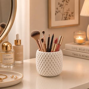 May include: A white cylindrical makeup brush holder with a textured diamond pattern containing various makeup brushes, pencils, and lipsticks. Nearby are a glass perfume bottle, a foundation bottle, a small dish with jewelry, and two stacked books.