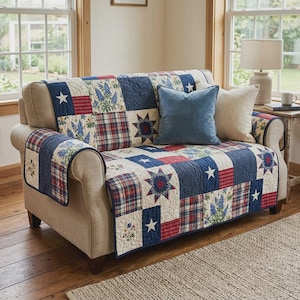 May include: A beige loveseat with a patchwork quilt featuring blue, red, and white squares. The quilt has star, floral, and plaid patterns. Two throw pillows, one blue and one beige, rest on the loveseat. A lamp and side table are visible.