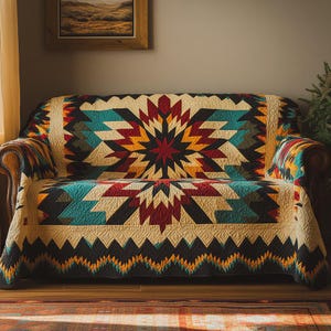 May include: A sofa covered with a colorful quilt featuring a geometric starburst design. The quilt incorporates teal, red, yellow, and black patterns. The sofa has brown leather arms and is placed in a room with a painting and a small tree.