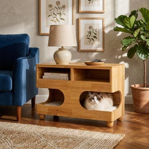 May include: A light-colored wooden pet house and side table featuring two upper open shelves and two lower arched openings. A fluffy cat with white and grey fur rests inside one of the lower compartments. The unit stands on four rounded feet.