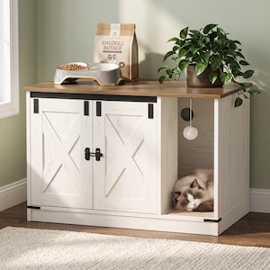 May include: A white wooden cat litter box enclosure with a brown top, featuring two sliding barn-style doors with black hardware, an open side compartment with a hanging toy, a cat food bowl, and a bag of Ragdoll Royale cat food on top.