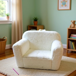 May include: A small, white, plush armchair sits on a cream-coloured rug in a child's room. The chair has a rounded back and armrests. A pink pencil lies on the rug. A framed picture of a teddy bear hangs on the wall.