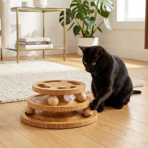 Peut inclure: Un chat noir interagit avec un jouet pour chat en bois à trois niveaux, doté de plateformes circulaires et de boules en bois lisses. Une petite balle blanche perforée est visible sur le niveau intermédiaire. Le jouet est en bois clair naturel.