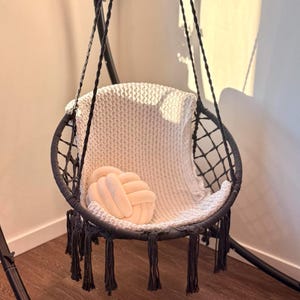 May include: A round hanging chair with a black frame and a woven seat. A cream blanket and a light peach knot cushion are inside. The chair has black fringe and is suspended from a black metal frame.