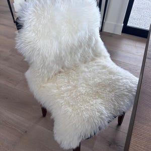 May include: A white, fluffy sheepskin chair cover. The cover is draped over a dark-colored chair with wooden legs. The sheepskin has a thick, shaggy texture, creating a soft and inviting appearance. The chair is positioned in a well-lit room.
