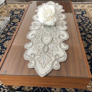 May include: A decorative beaded table runner in ivory and silver, featuring an intricate floral design. The runner is placed on a wooden coffee table with a glass top, and a vase of white roses sits atop it. The table is set on a patterned rug.