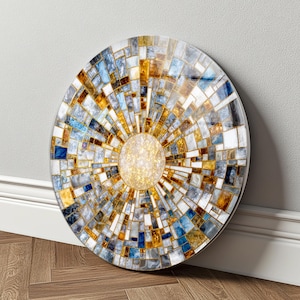 Op de afbeelding: Een cirkelvormig wandkunstobject met een mozaïekpatroon van rechthoekige tegels in goud-, blauw-, wit- en grijstinten. Het ontwerp straalt uit vanuit een centraal, getextureerd goudkleurig cirkelvormig middelpunt met een zonnestraaleffect.