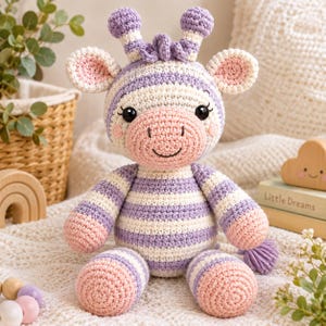 Peut inclure: Une peluche au crochet en forme de girafe avec un motif rayé lavande et crème. Le jouet présente un museau rose tendre, deux petites cornes sur le dessus de la tête, de grandes oreilles rondes et des yeux noirs avec des cils brodés.