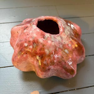 Op de afbeelding: Een handgemaakte keramische vaas in koraalroze met een donkerbruin interieur. De vaas heeft een onregelmatige, organische vorm met een centrale opening. Het oppervlak heeft gespikkelde details en een glanzende afwerking. Een decoratief kunstobject.