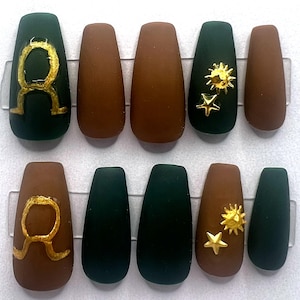 May include: A set of ten matte finish, coffin-shaped false nails. The nails are a mix of dark green and brown. Some nails feature gold accents, including a Taurus symbol, a sun, and stars.