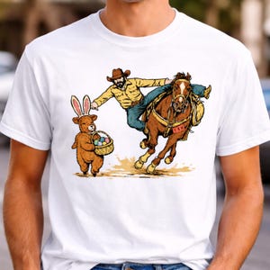 May include: White t-shirt featuring a humorous illustration of a cowboy on a bucking horse, a teddy bear with bunny ears holding a basket of Easter eggs. The cowboy is wearing a yellow shirt and blue jeans.