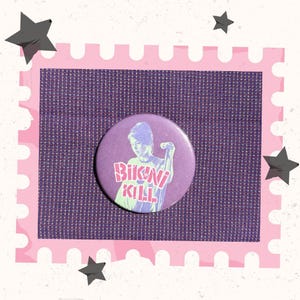 Puede incluir: Un botón redondo morado con las palabras "BIKINI KILL" en una fuente estilizada. El botón presenta una ilustración en blanco y negro de una persona sosteniendo un micrófono. El botón está sobre un fondo de tela morada.