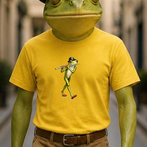 Könnte beinhalten: Eine Person mit einem Froschkopf trägt einen schwarzen Bowler und ein gelbes T-Shirt. Das T-Shirt zeigt einen Cartoon-Frosch, der Flöte spielt. Die Person trägt auch eine braune Hose und einen braunen Gürtel.