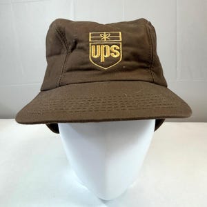 May include: A brown bucket hat featuring the gold embroidered UPS logo on the front. The hat has a structured crown and a wide, downward-sloping brim with visible stitching detail. The fabric appears to be a durable cotton twill material.