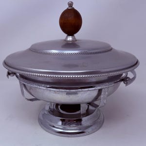 May include: A silver-toned metal chafing dish with a round base, a bowl-shaped body, and a matching lid. The lid features a dark brown wooden spherical knob at the top and a decorative beaded edge. The dish includes two side handles and a stand.