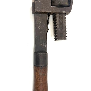 May include: A vintage pipe wrench featuring a dark, weathered metal head with a serrated jaw and an adjustable knurled nut mechanism. The tool has a long, straight metal shank connected to a smooth, dark brown wooden handle with a rounded end.