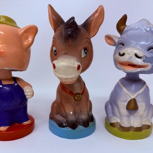 Könnte beinhalten: Drei Wackelkopf-Figuren bestehend aus einem rosa Schwein in blauer Latzhose mit gelbem Hut, einem braunen Esel mit rotem Halsband und einer hellvioletten Kuh mit weißer Glockenkette. Jede Figur steht auf einem runden Sockel in Rot, Blau oder Grün.