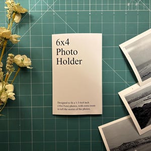 May include: A white 6x4 Photo Holder with text, designed for 10x15cm photos. The holder is on a green grid surface, with black and white photos and dried flowers nearby.