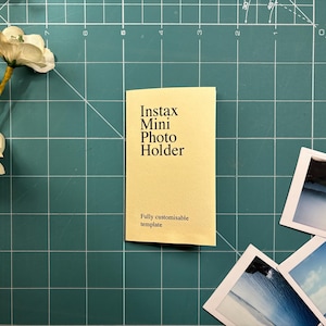 Puede incluir: Un portafotos Instax Mini amarillo con el texto "Instax Mini Photo Holder" y "Fully customisable template" sobre una esterilla de corte verde. Varias fotos Instax están esparcidas alrededor del soporte, con flores blancas en la esquina superior izquierda.