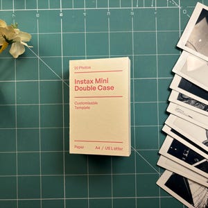 以下が含まれることがあります： 赤い文字で「20 Photos」と書かれたクリーム色のInstax Mini Double Caseが、緑色のカッティングマットの上に置かれています。Instaxの写真の束と白い花の枝も写っています。ケースには「Customisable Template」と書かれています。