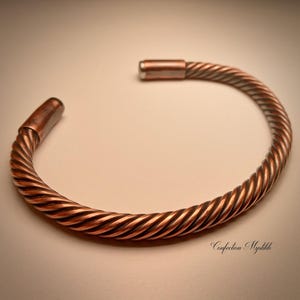 May include: A copper-coloured, twisted metal bracelet. The bracelet has a spiral design and is open-ended. The ends of the bracelet are capped with a solid copper piece. The bracelet is a jewelry item.