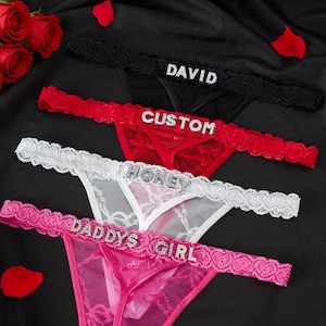 May include: Four thong underwear in black, red, white, and pink, each with a different word spelled out in rhinestones: "DAVID", "CUSTOM", "HONEY", and "DADDY'S GIRL". The underwear is displayed on a black surface with red rose petals and a bouquet of red roses.