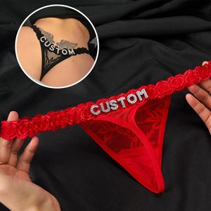 Può includere: Perizoma in pizzo rosso con la scritta "CUSTOM" in strass argentati. La fascia in vita presenta un bordo a forma di cuore. Un secondo perizoma in pizzo nero con "CUSTOM" è visibile sullo sfondo.