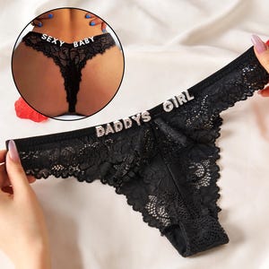 May include: Black lace thong underwear featuring silver rhinestone lettering that spells out DADDYS GIRL on the front waistband and SEXY BABY on the back waistband, shown in a circular inset image.