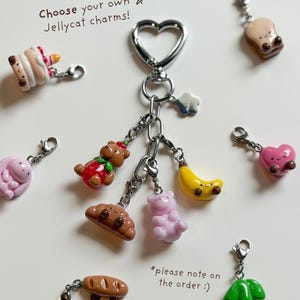 May include: A collection of Jellycat charms, including a cake, bear, croissant, banana, pink bear, bread, heart, and cherries. The charms are attached to a silver heart-shaped keychain. Text on the image reads "Choose your own Jellycat charms!"