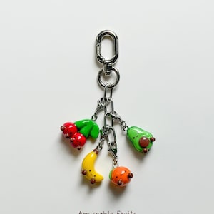 May include: A silver keychain with a lobster clasp holding charms shaped like a cherry, avocado, banana, and orange. The charms are colorful and have smiling faces. The text "Amuseable Fruits" is at the bottom.